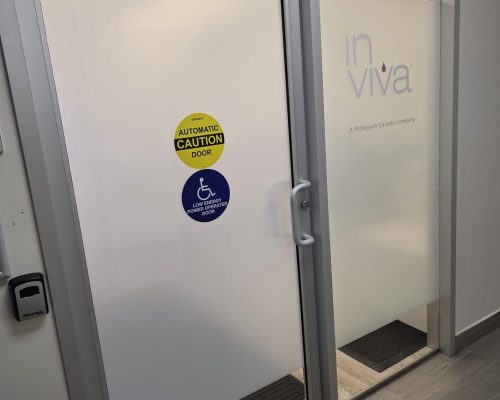 A frosted glass office door with in viva and A McKesson Canada Company printed on it. Yellow and blue stickers indicate the door is automatic and accessible for people with disabilities.
