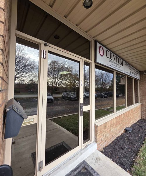 A glass door entrance to an office building with the CENTUM logo and name on the window. Reflections of trees, cars, and a cloudy sky are visible in the glass. A black mailbox is mounted on the brick wall.