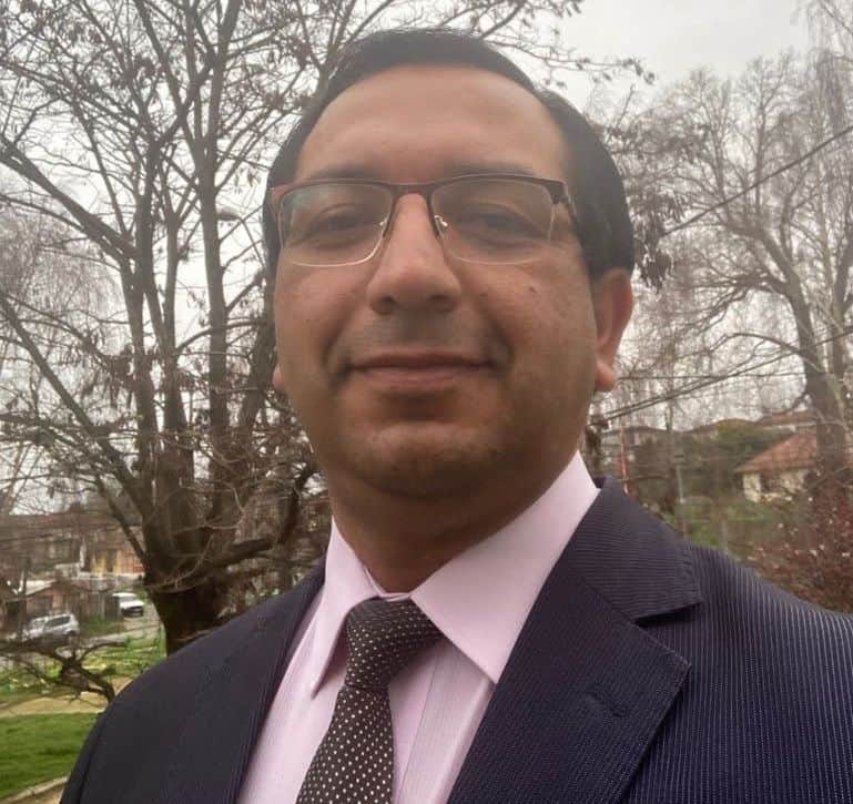 Filósofo en exteriores con árboles y casas, vestimenta formal, gafas y sonrisa, en un día nublado.