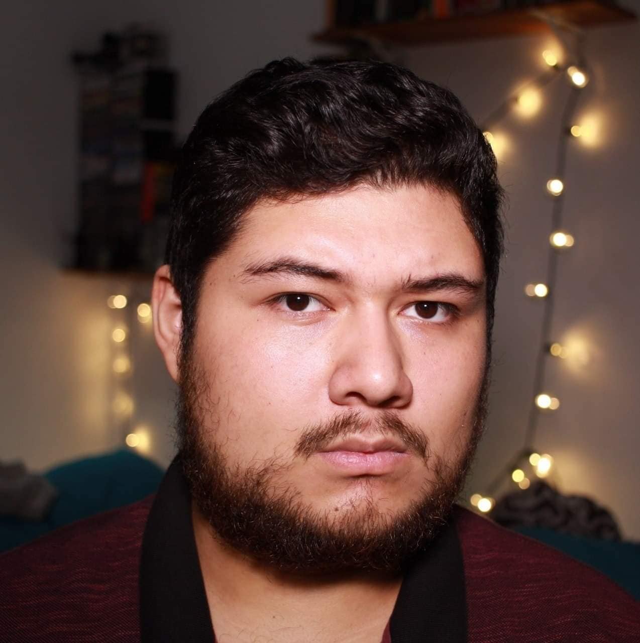 Joven con barba y cabello oscuro, retrato en interiores con luces decorativas en el fondo.