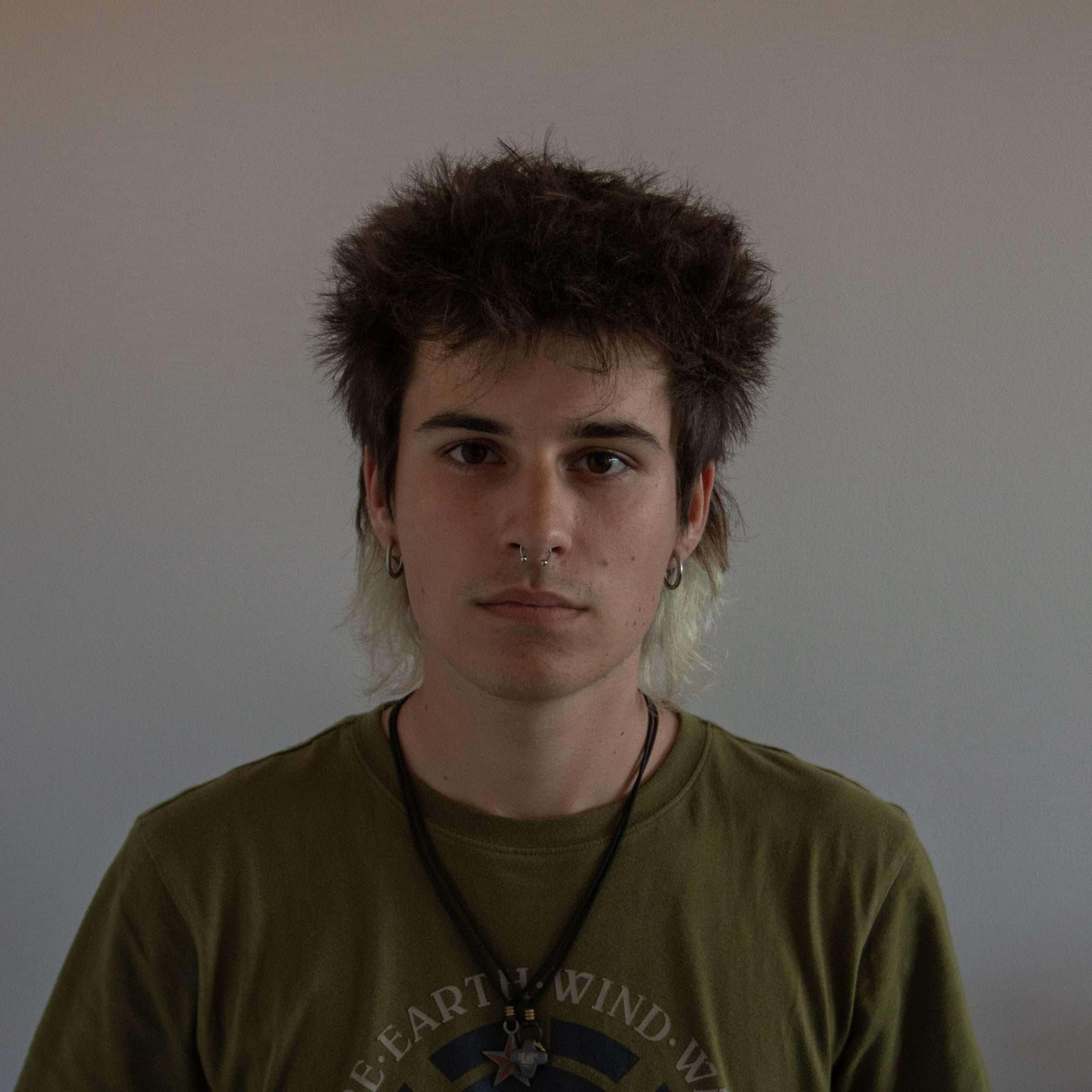 Joven con estilo punk, cabello desordenado y piercing en la nariz, en fondo gris neutro.