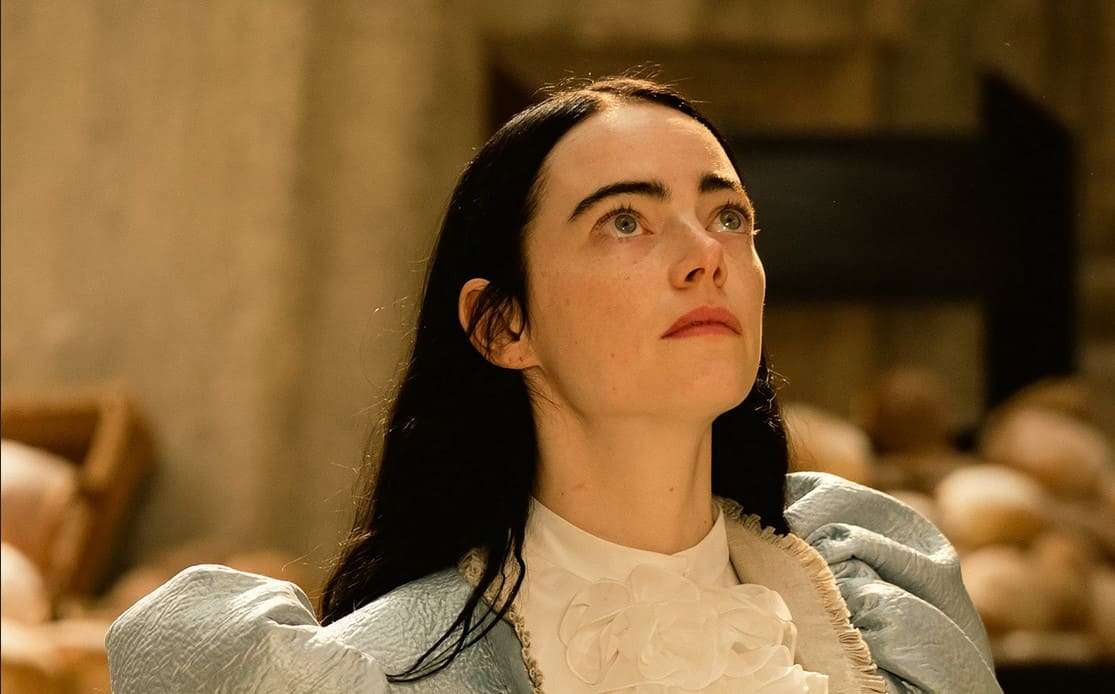 Una mujer con cabello oscuro y vestido elegante en un escenario teatral con fondo de ladrillos.