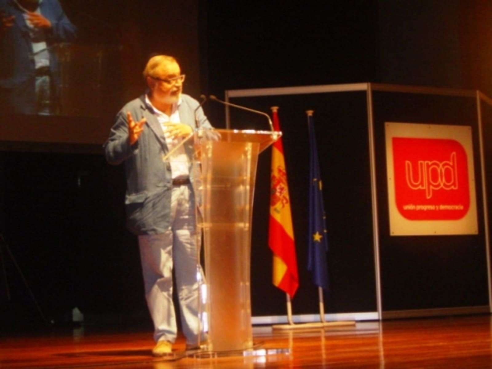 Pensador en conferencia con banderas de España y la Unión Europea en fondo oscuro.
