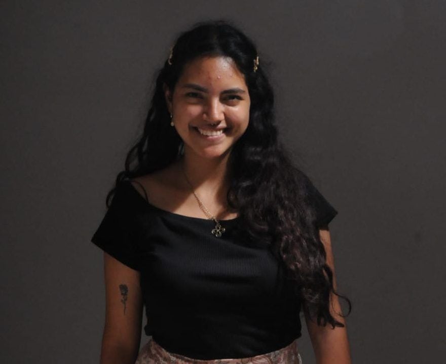 Joven sonriendo con cabello oscuro y accesorios, en fondo gris oscuro.