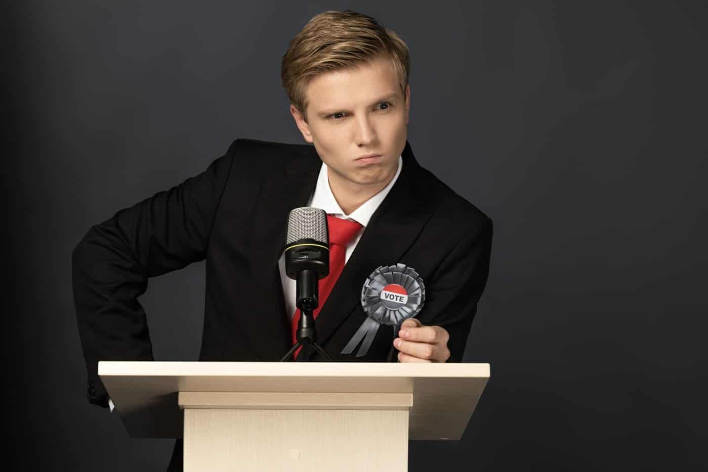 Político joven con expresión pensativa en un debate, vestido con traje y corbata roja, sosteniendo un símbolo de voto.