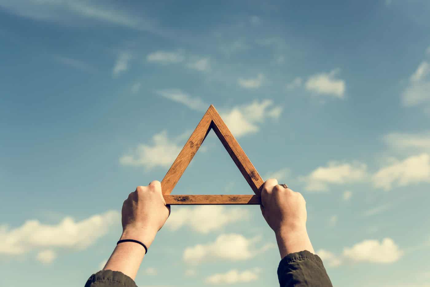 Triángulo de madera en manos contra cielo con nubes, símbolo de equilibrio filosófico y pensamiento.