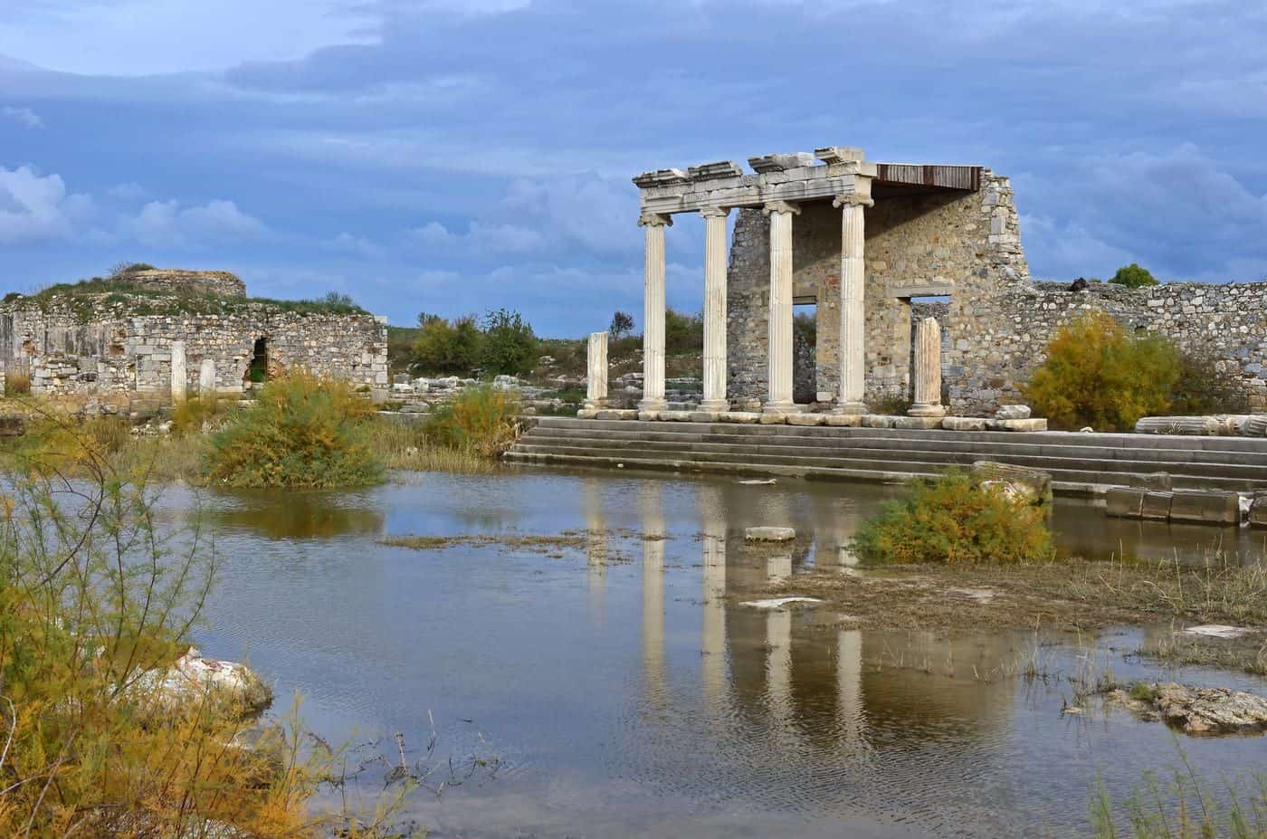 Templo greco en ruinas junto a un río, con columnas clásicas y paisaje natural.