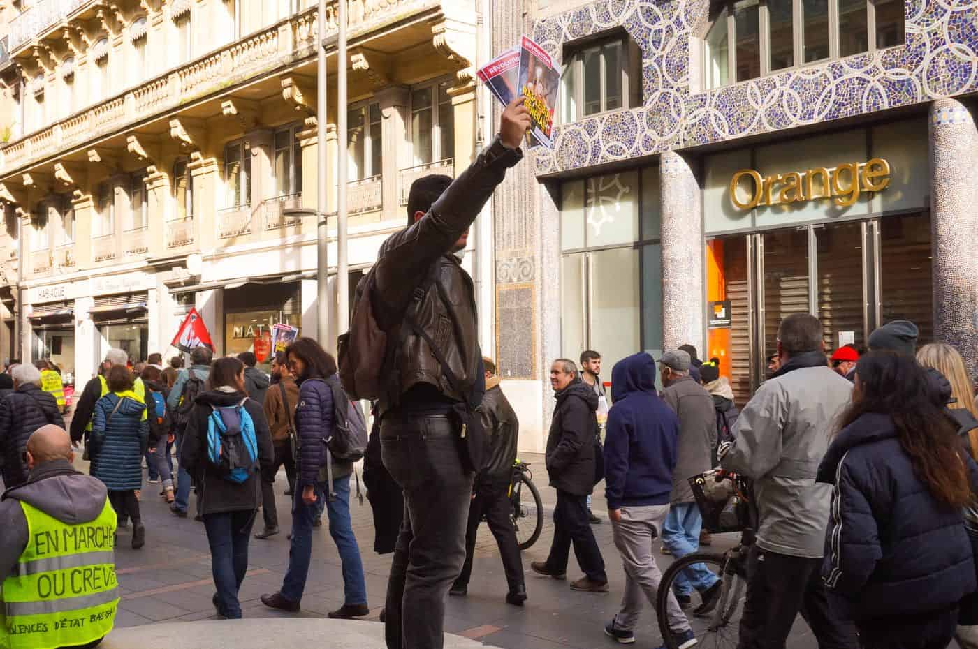 Manifestantes en la calle protestando, algunos con banderas y pancartas, en frente de un edificio con decoración artística en París.