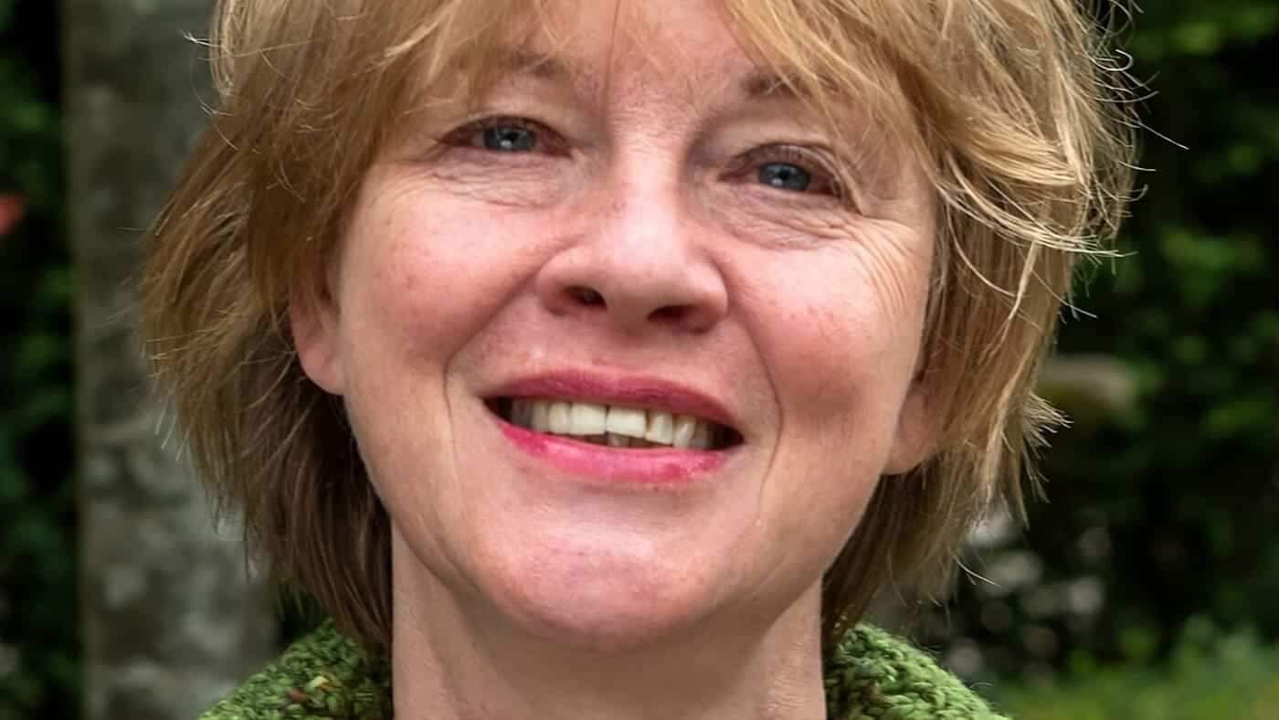 Retrato de una mujer mayor con cabello rubio y sonrisa, al aire libre con vegetación de fondo.