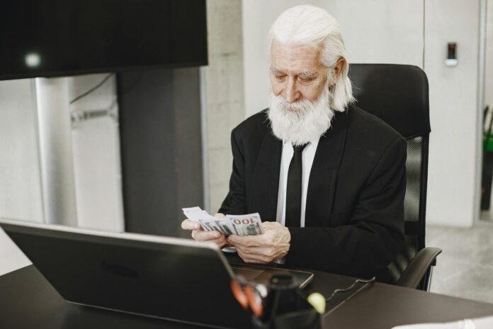 Senior businessman in suit counting money at office desk with a laptop. Elegance and financial focus