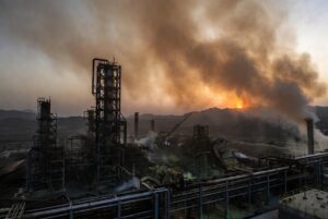 Massive LNG processing facility with damaged industrial towers, smoke rising from a petrochemical complex at dawn, orange and grey sky, heat distortion