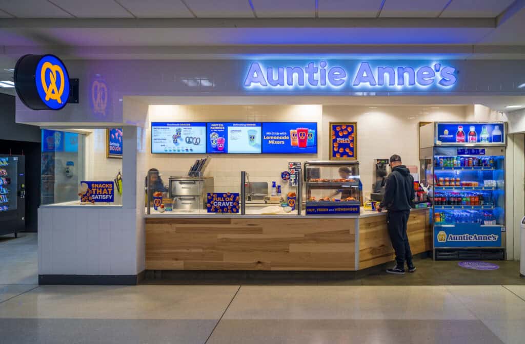 The Auntie Anne’s pretzel shop at John Glenn Columbus International Airport, featuring a bright illuminated sign and a counter displaying pretzels and snacks. A customer stands at the register, and a cooler filled with bottled drinks sits to the right. Digital menu boards above the counter show pretzel options and beverages.