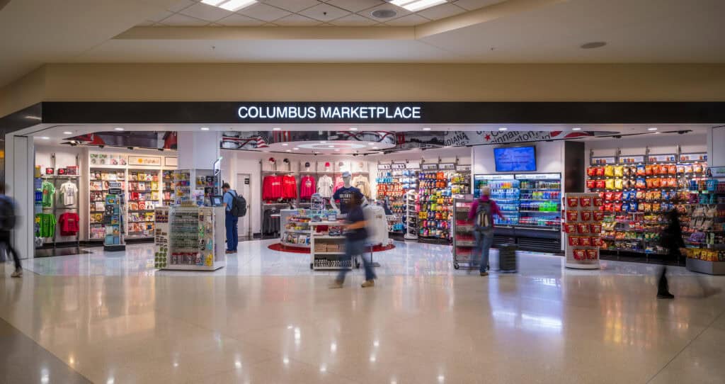 The Columbus Marketplace store at John Glenn Columbus International Airport, brightly lit with shelves of snacks, drinks, magazines, souvenirs, and Ohio-themed apparel. Several travelers browse inside while others walk past in a motion blur. The store’s name appears in large illuminated letters above the entrance.