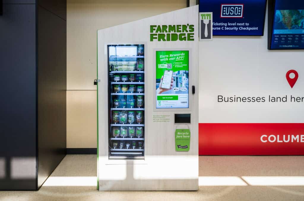 A Farmer’s Fridge vending machine at John Glenn Columbus International Airport, styled in light wood with green accents. The machine displays rows of fresh grab-and-go items like salads, bowls, and snacks in clear jars. A touchscreen on the right promotes the Farmer’s Fridge app and payment options. A small slot below the screen is labeled “Recycle jars here.” Bright sunlight casts shadows across the floor.