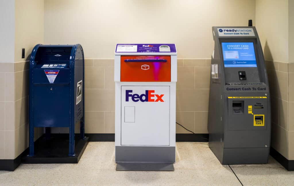 A USPS blue mail drop box, a white and orange FedEx drop box, and a gray ReadyStation machine for converting cash to a prepaid card are lined up against a beige tiled wall at John Glenn Columbus International Airport.