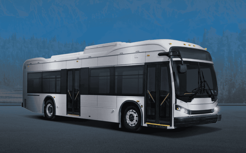 A silver transit bus with its front doors open, parked on asphalt with blurred blue mountains and trees in the background.