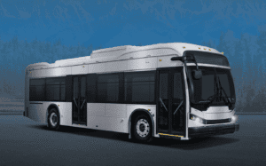 A silver transit bus with its front doors open, parked on asphalt with blurred blue mountains and trees in the background.