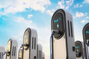 Multiple modern electric vehicle charging stations with glowing blue screens and cables plugged in, set against a blue sky with white clouds.