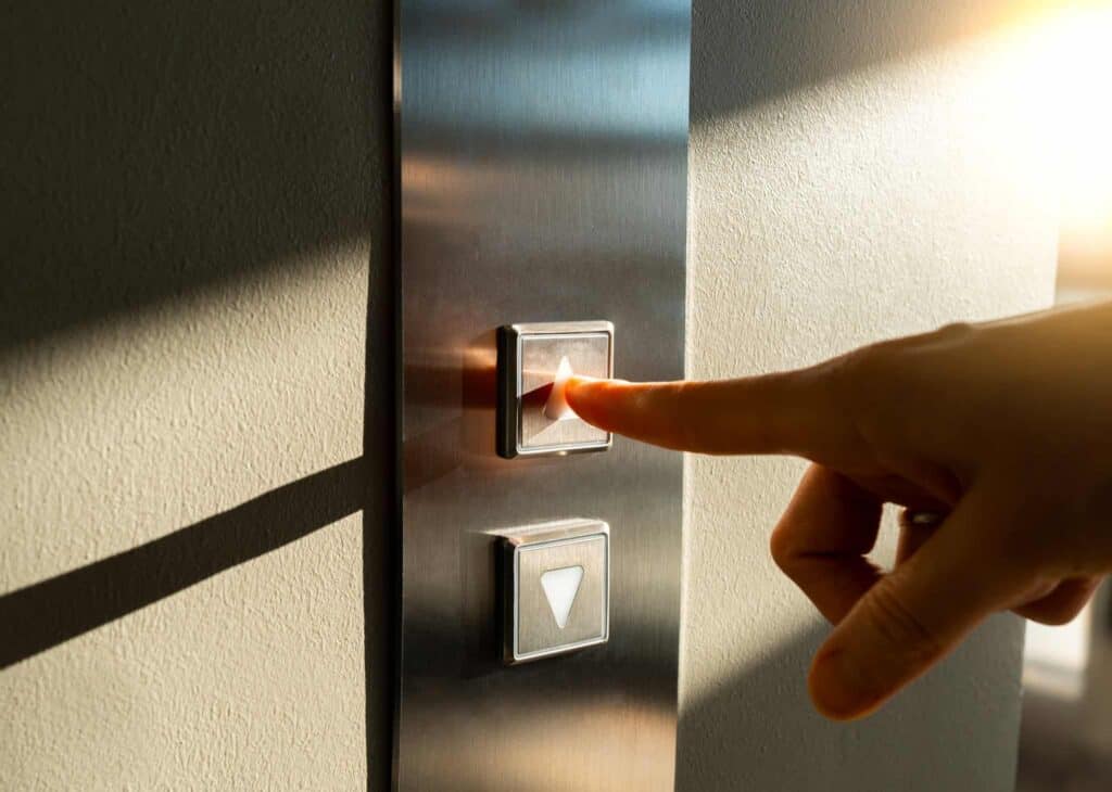 A hand pressing an illuminated 'up' elevator button on a brushed metal panel, with a 'down' button below it.