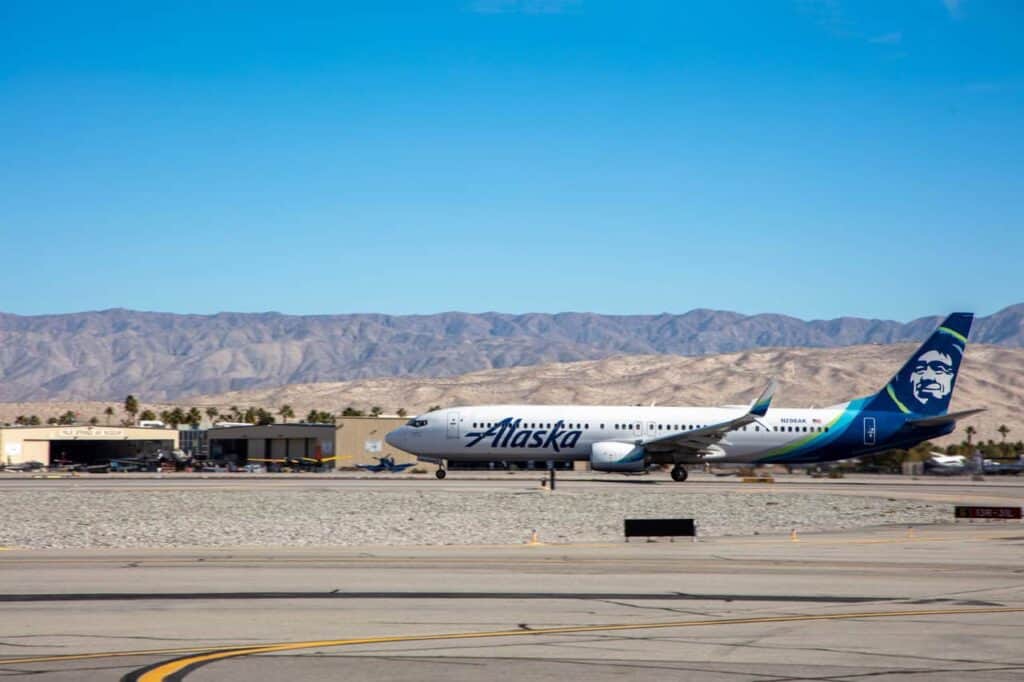 An Alaska Airlines Boeing 737 aircraft on an airport tarmac with mountains and a clear blue sky in the background.