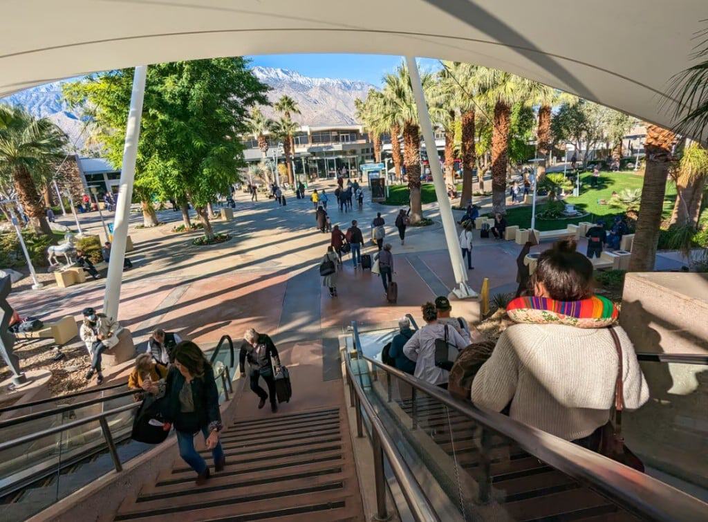 View from above of people descending stairs and an escalator into a sunny outdoor plaza with palm trees, a modern building, and distant snow-capped mountains.