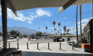 View from under a concrete overhang showing a parking lot with cars, palm trees, and a police SUV, with mountains and a blue sky in the background.