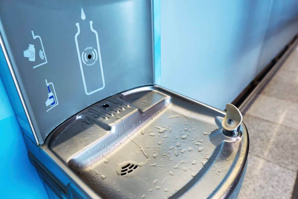 A stainless steel water fountain and bottle filling station with water droplets, featuring a sensor-activated bottle filler and a traditional drinking spout. Instructional graphics are visible on the back panel.
