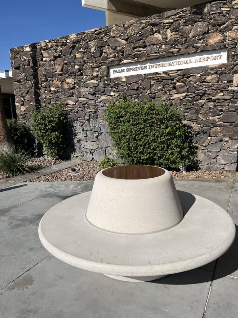 A white sign for Palm Springs International Airport on a dark stone wall, next to green bushes and a modern concrete planter.