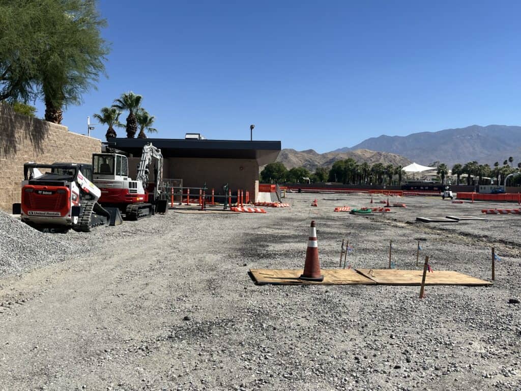 A construction site with a red and white Bobcat skid-steer loader and a Takeuchi mini excavator on gravel, surrounded by construction barriers, with palm trees and arid mountains in the background under a clear blue sky.