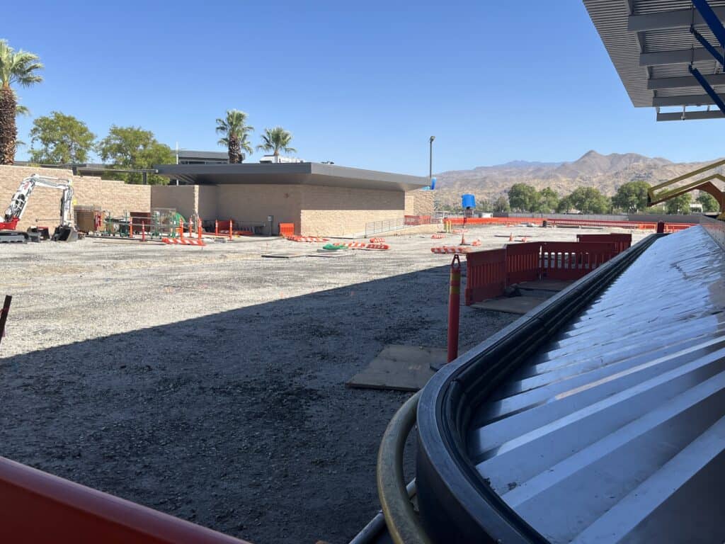 An outdoor construction site with gravel ground, an excavator, orange barriers, a building, palm trees, and distant mountains under a clear blue sky.