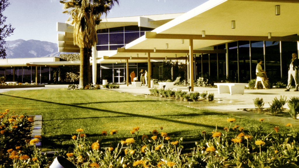 Mid-century modern building with large windows and overhanging roof, surrounded by green lawn, palm trees, and flowers, with mountains in the background.