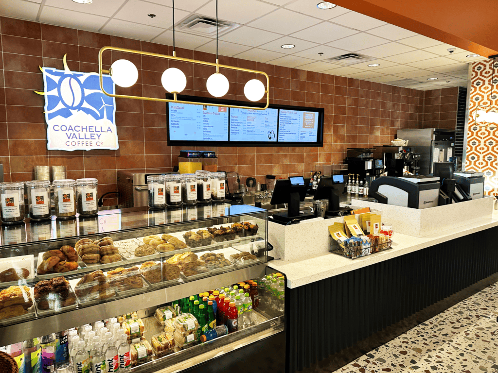Interior of Coachella Valley Coffee Co, showing a display case with pastries and drinks, a counter with coffee machines and POS systems, a menu board, and a blue 'Coachella Valley Coffee Co' sign.