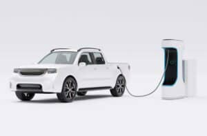 A white electric pickup truck is charging at a white and blue electric vehicle charging station against a white background.