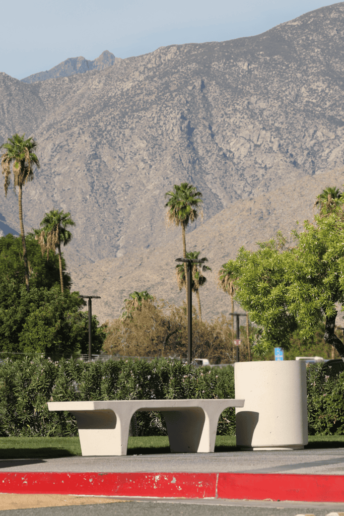 A concrete bench and planter in front of green bushes, palm trees, and rocky mountains under a blue sky.