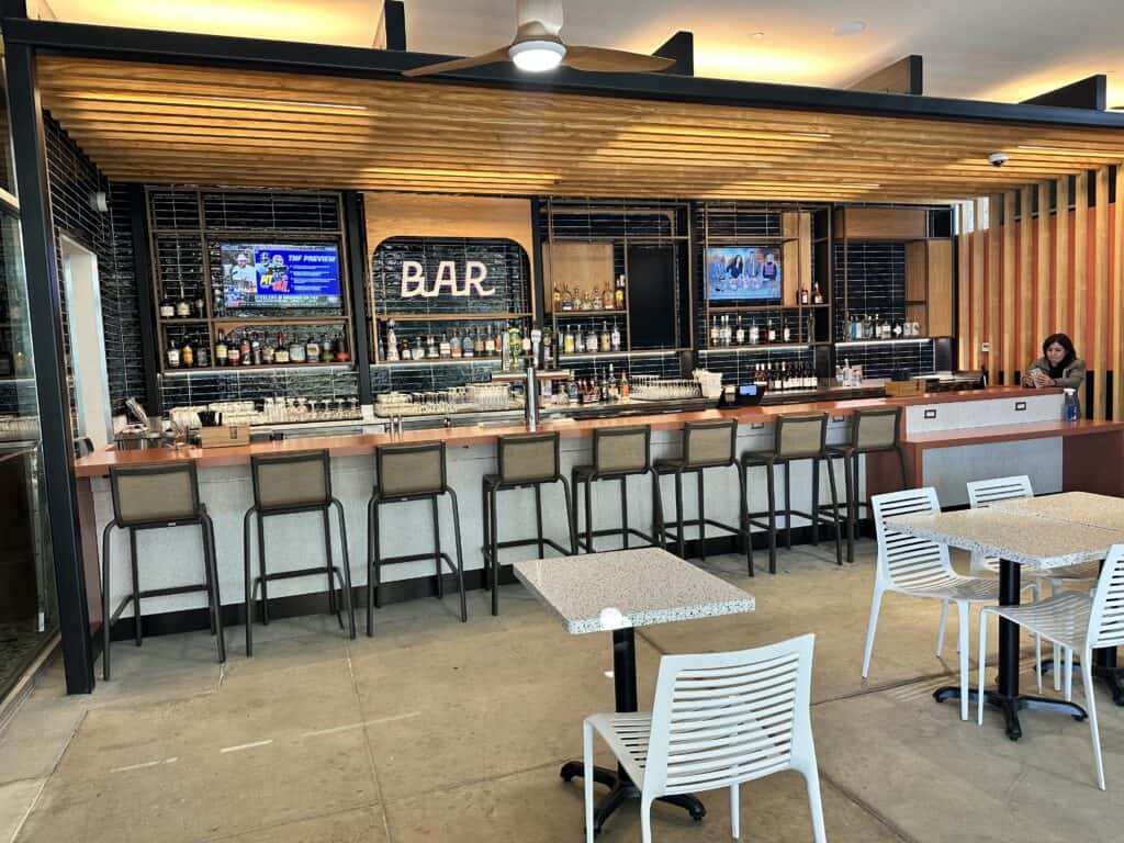 A modern bar area with a copper counter, black tiled backsplash, liquor bottles, two TVs, and a 'BAR' sign. Bar stools and white tables with chairs are in the foreground. A woman is on her phone at the end of the bar.