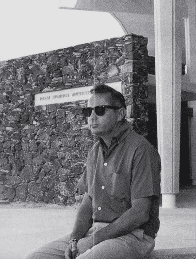 Black and white photo of a man in sunglasses and a short-sleeved shirt sitting in front of a stone wall with a "Palm Springs" sign.