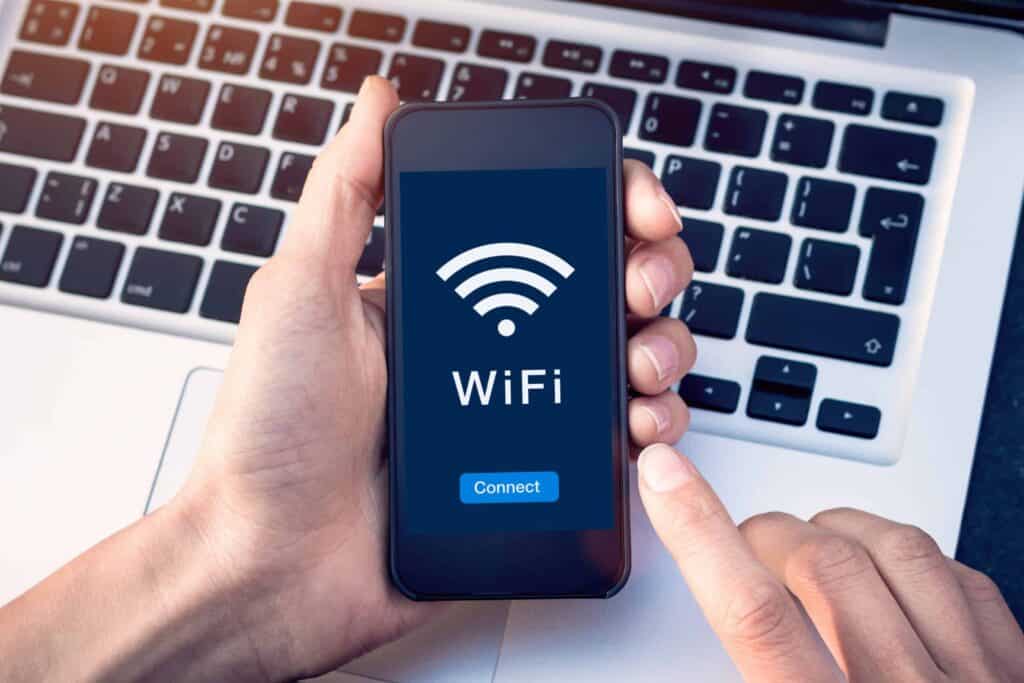 Hands holding a smartphone displaying a Wi-Fi icon, the word 'WiFi', and a 'Connect' button on a blue screen, with a laptop keyboard in the background.