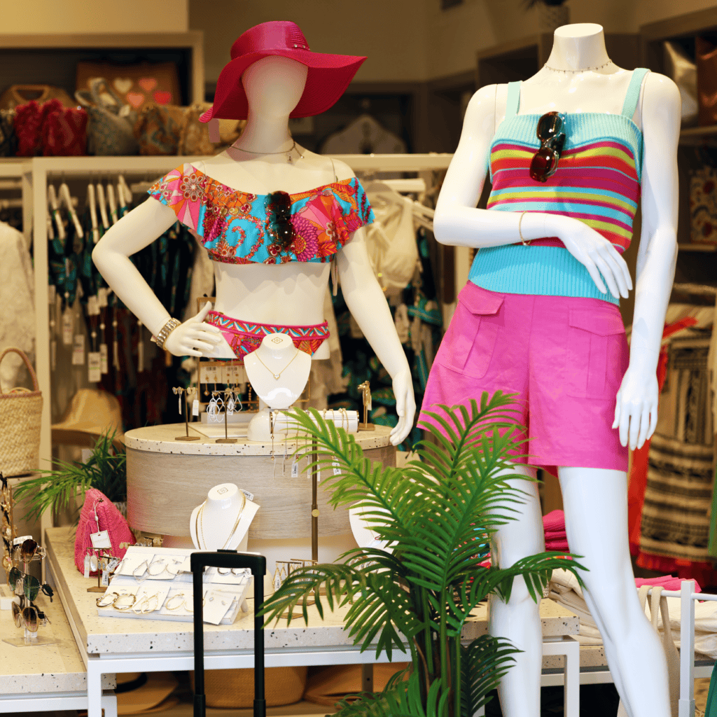 Two mannequins in a store display, one in a floral bikini and pink hat, the other in a striped top and pink shorts, surrounded by jewelry and tropical decor.