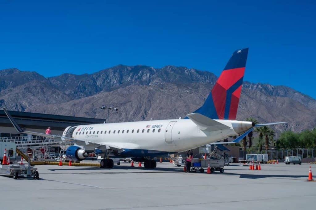 A Delta Connection airplane, white with blue and red tail, parked on a tarmac with mountains in the background. Ground crew and equipment are visible around the plane.