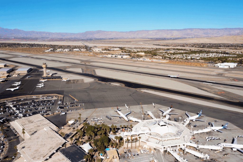 Aerial view of an airport with planes, runways, and terminal buildings, set against a desert landscape with mountains and wind turbines in the distance.