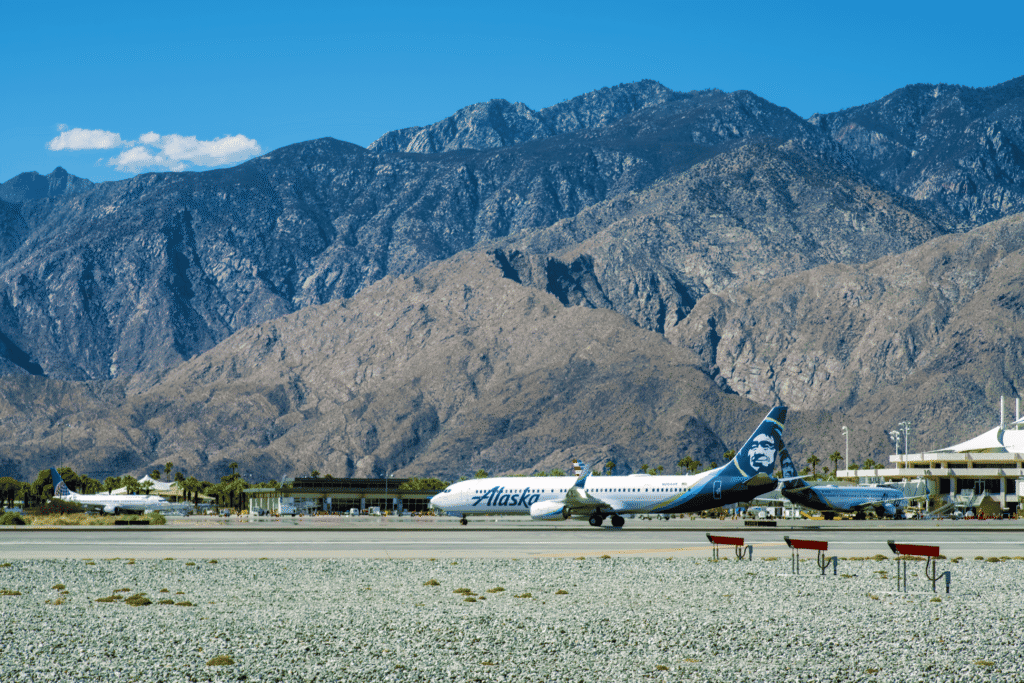 An Alaska Airlines plane on an airport tarmac with large, rocky mountains in the background under a blue sky.