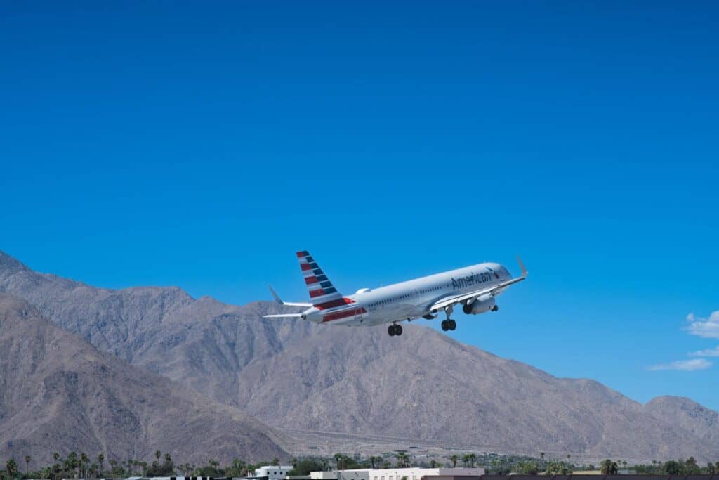 An American Airlines plane with landing gear down ascends into a clear blue sky, with arid mountains and palm trees in the background.