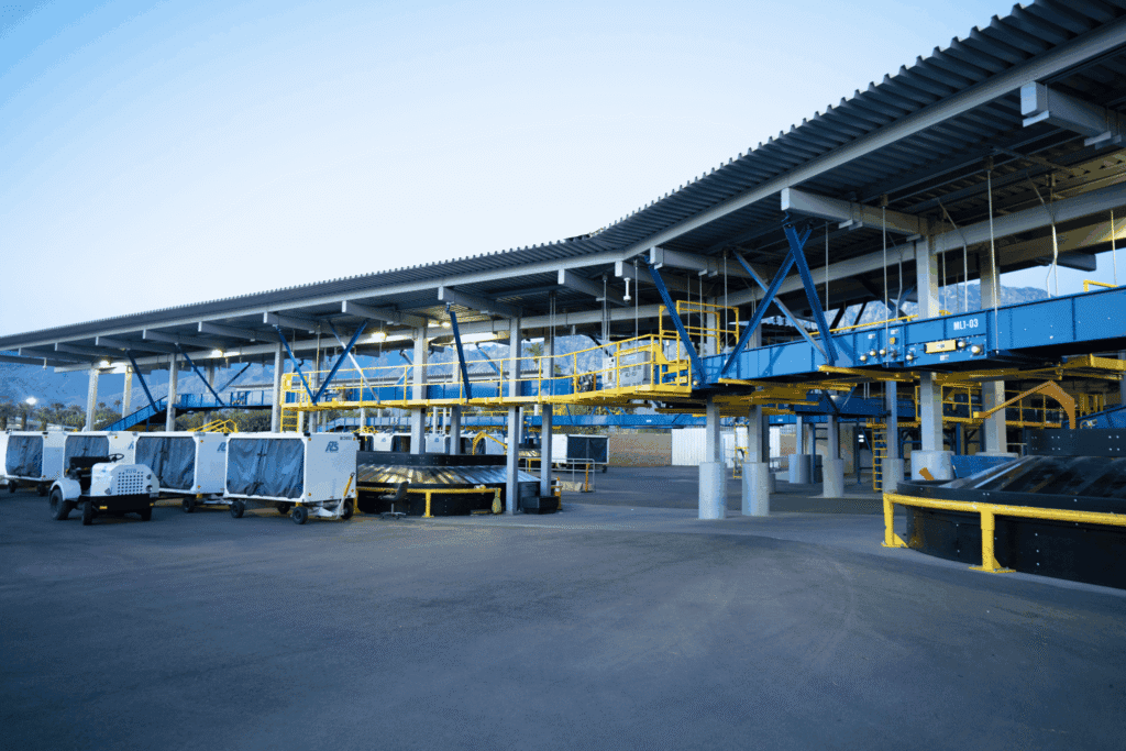 An outdoor baggage handling facility with blue and yellow conveyor belts, a white tug pulling baggage carts, and mountains in the background.