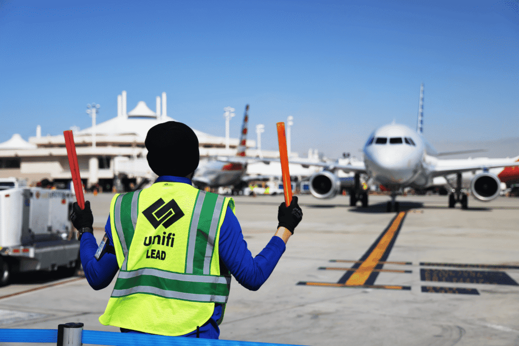 A ground crew member in a high-visibility vest and black beanie stands on an airport tarmac, holding two orange marshaling wands, facing a parked passenger airplane.