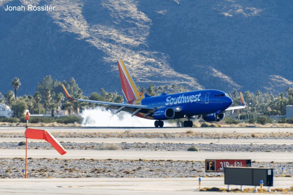 A blue Southwest Airlines Boeing 737 airplane landing on a runway, with smoke rising from its wheels. Mountains and palm trees are in the background.