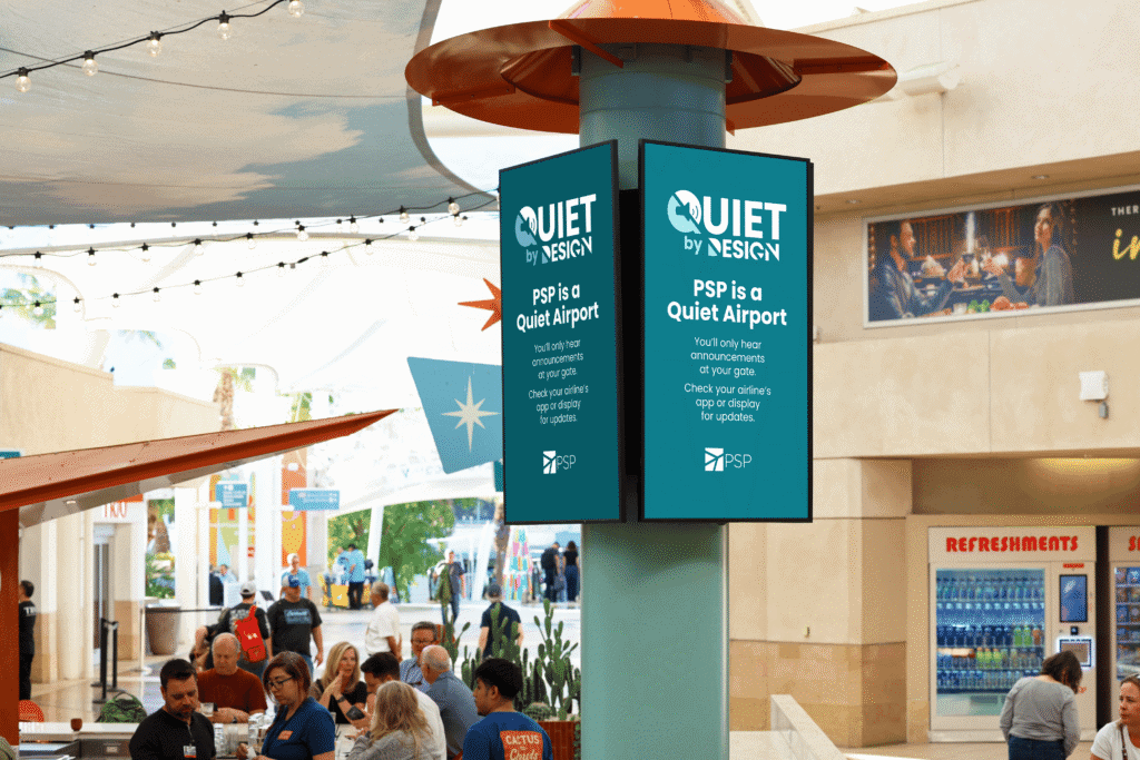 Two digital screens on a pillar in an airport terminal display 'PSP is a Quiet Airport' with instructions to check apps for updates. People are visible in the foreground and background.