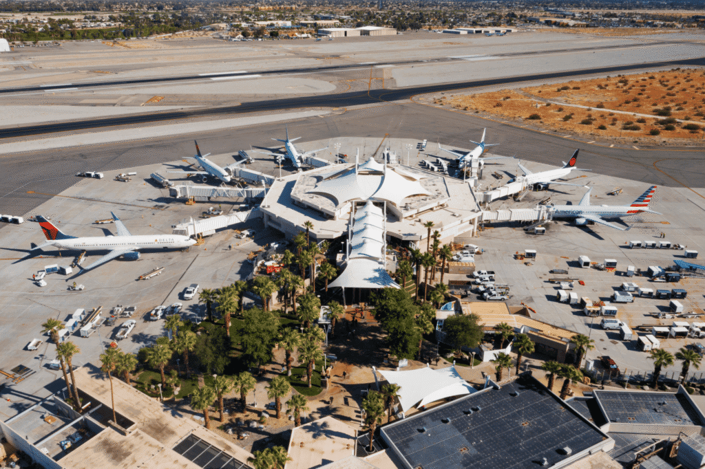 Aerial view of an airport terminal with several commercial airplanes, including Delta, American, and Air Canada, parked at gates. The terminal features white canopies and is surrounded by palm trees and an arid landscape.
