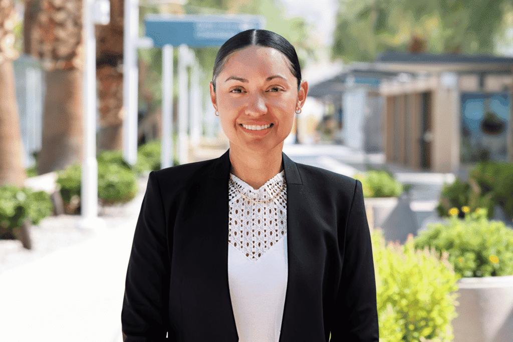 A smiling woman with dark hair in a black blazer and white lace top, standing outdoors.