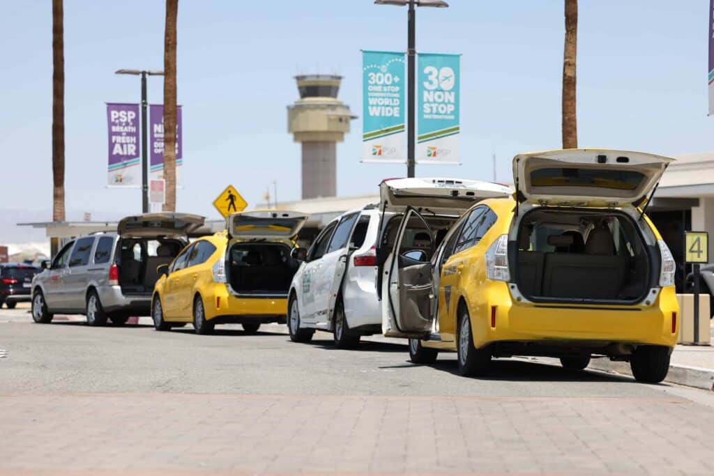 A line of yellow and white taxis with open trunks parked at an airport, with an air traffic control tower and promotional banners in the background.