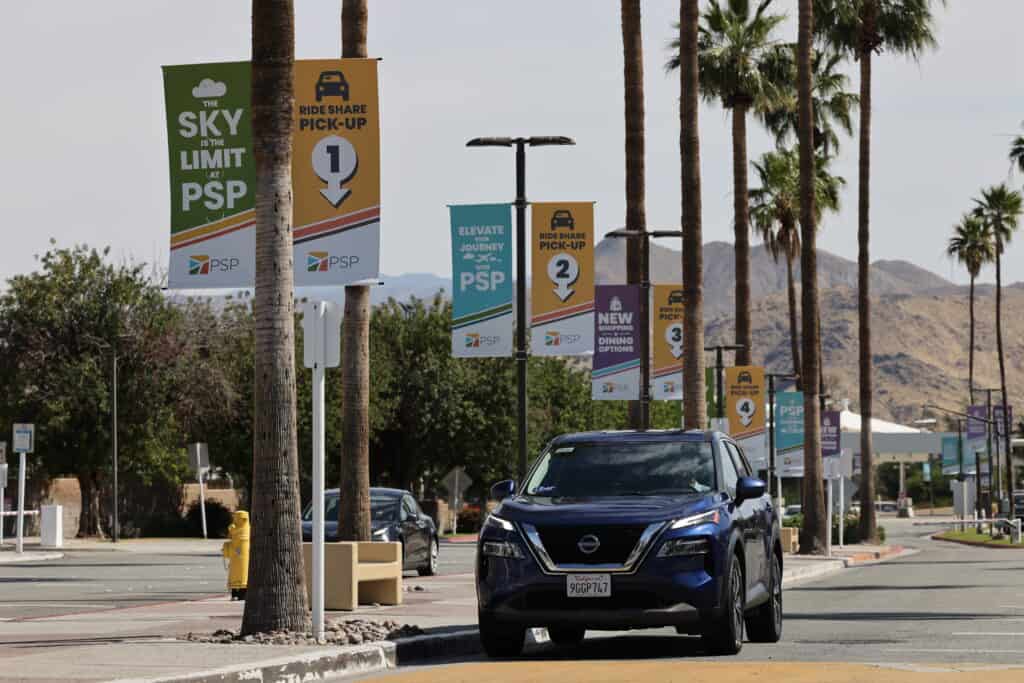 A blue Nissan SUV with an Uber sticker is parked on a road lined with palm trees and colorful banners for PSP airport ride-share pick-up zones, with mountains in the background.